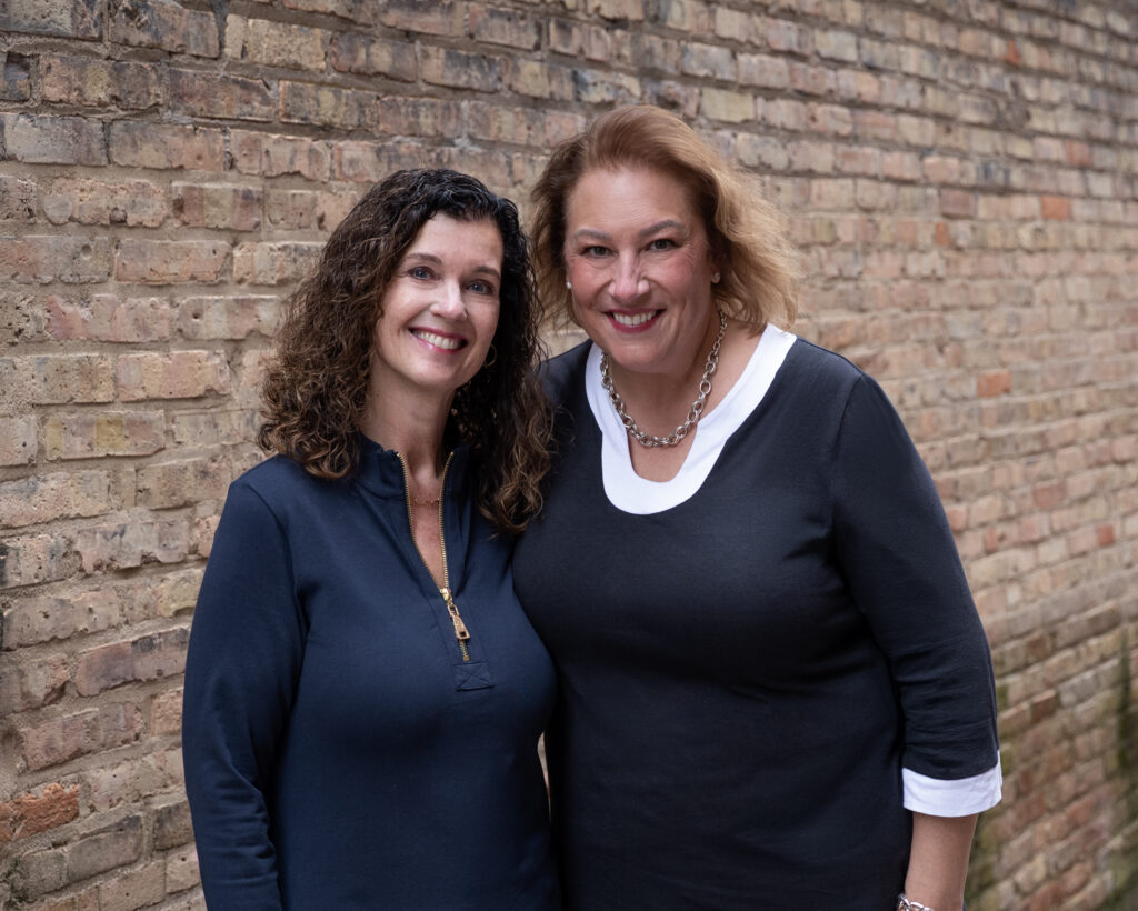 Beth Theile & Amanda Burnham posing in front of an exterior brick wall.